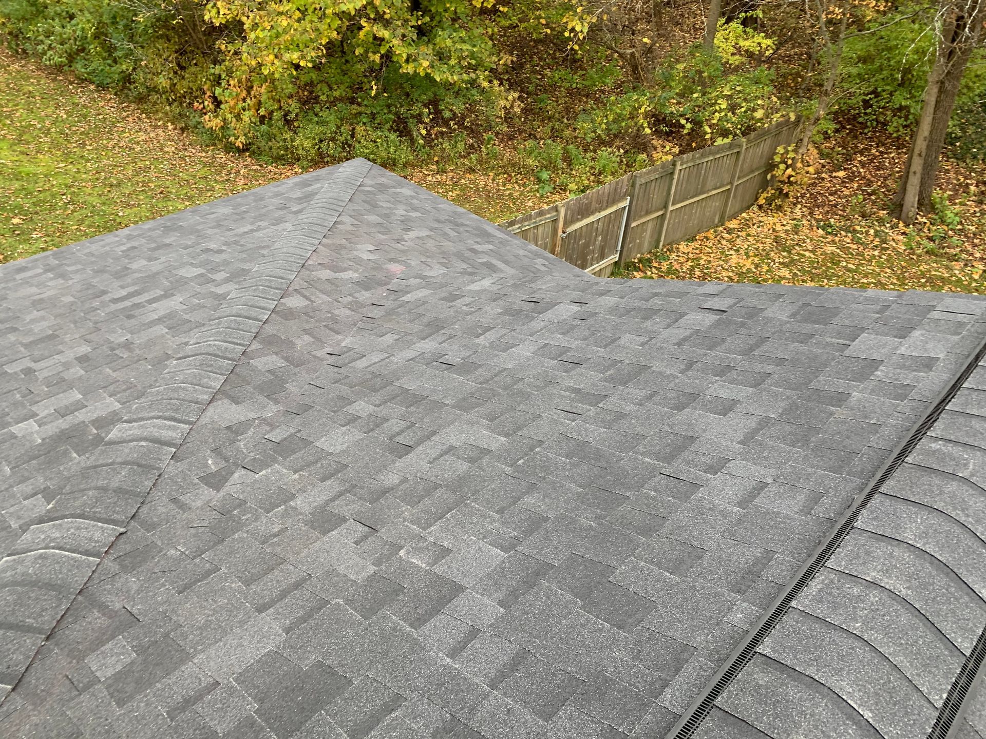 Gray shingled roof corner beside a fenced yard with autumn foliage below.