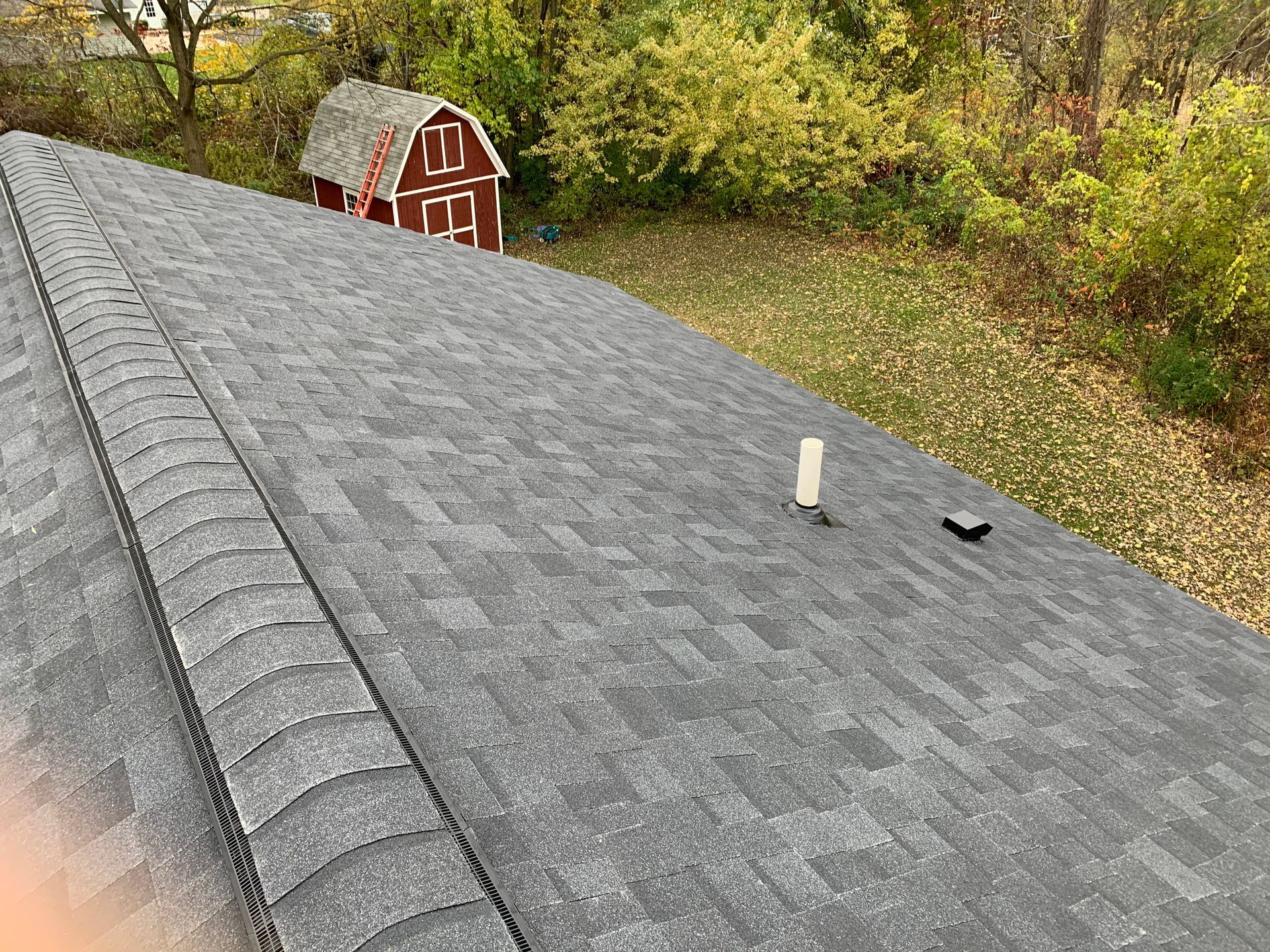 Gray shingled roof with vent pipes, overlooking a small red barn and autumn trees.