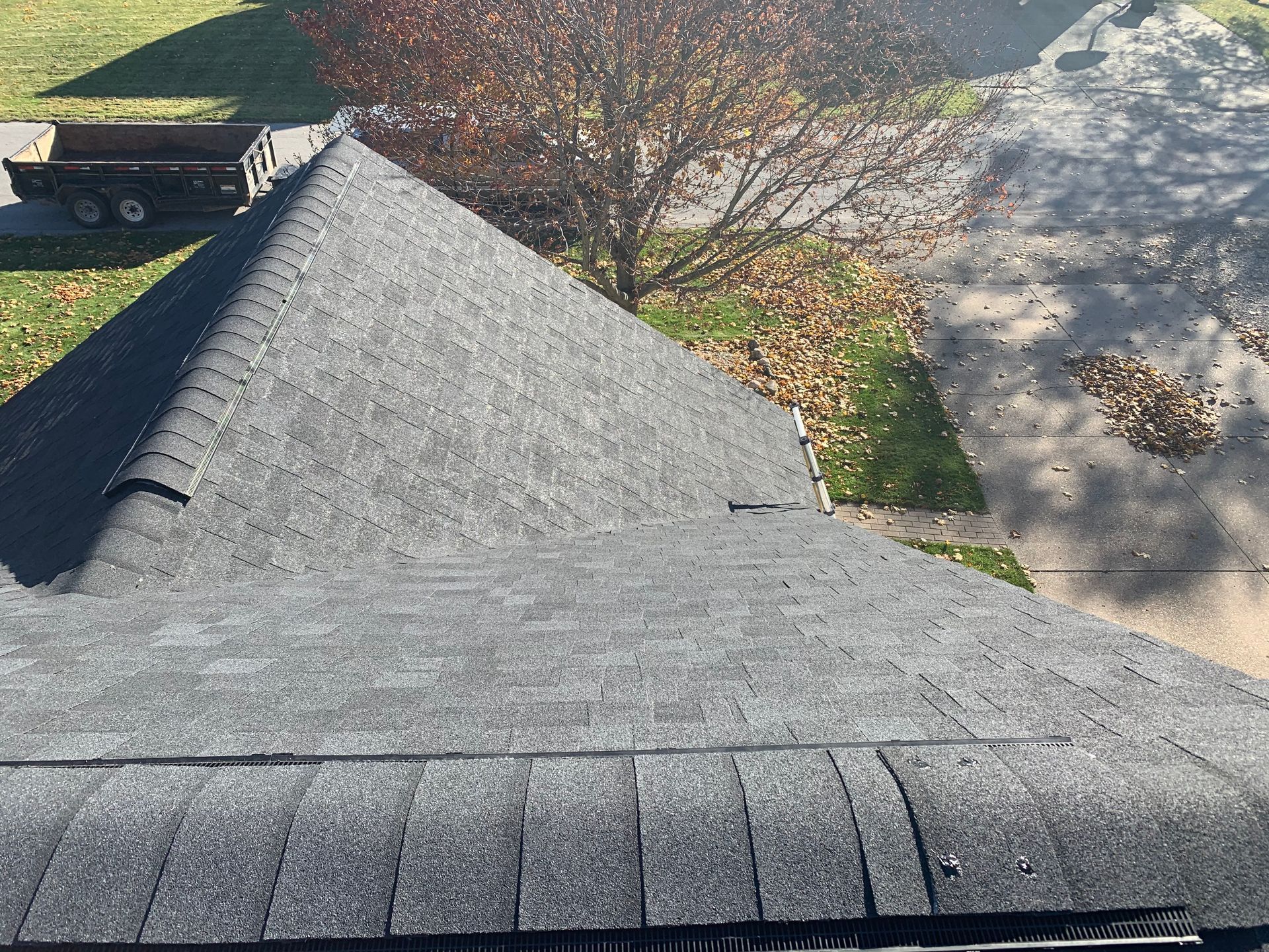 Gray shingled roof with a steep ridge, seen from above beside a driveway and tree shadows.