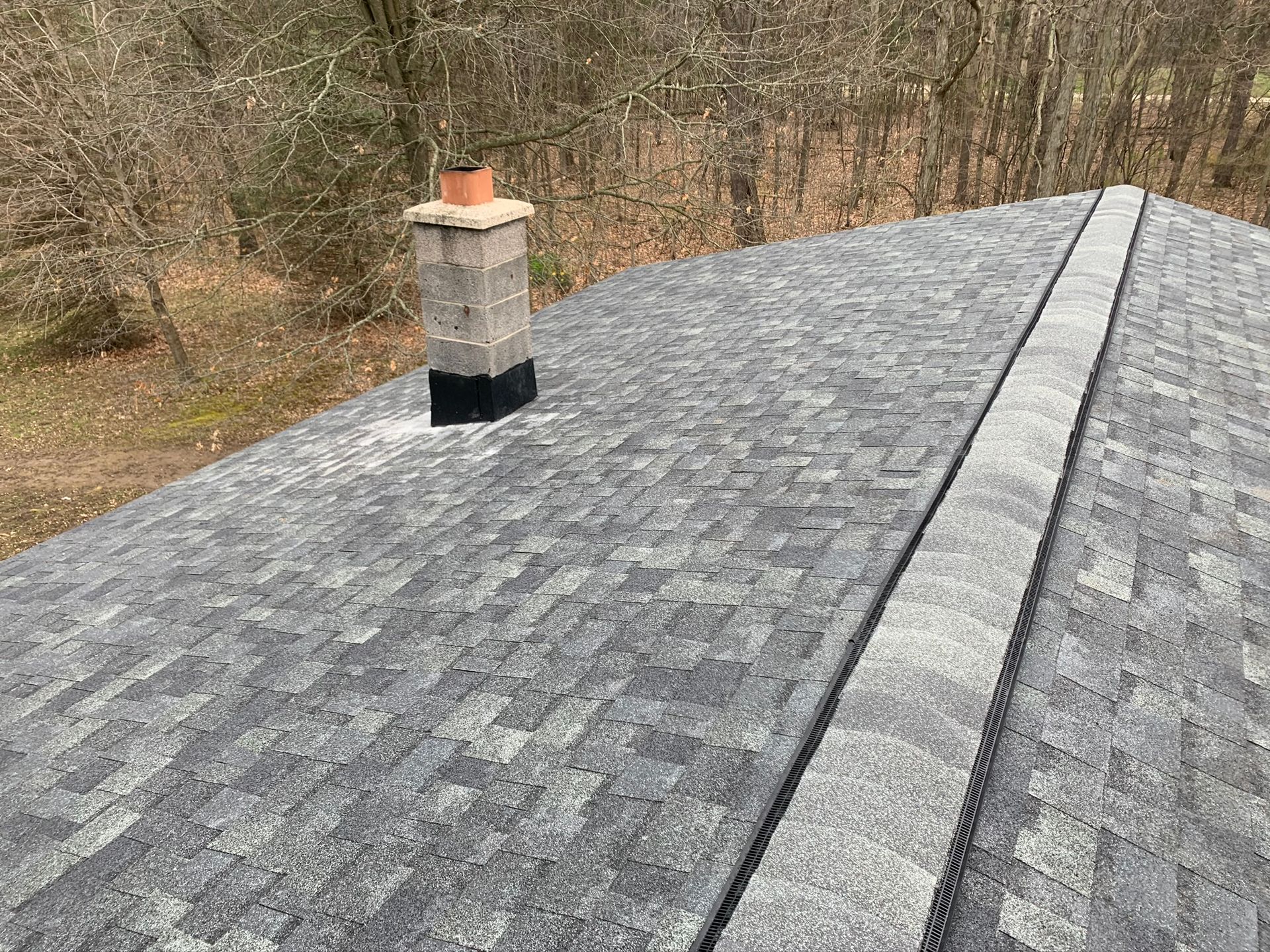 Gray shingled roof with a brick chimney, viewed from above beside leafless trees.