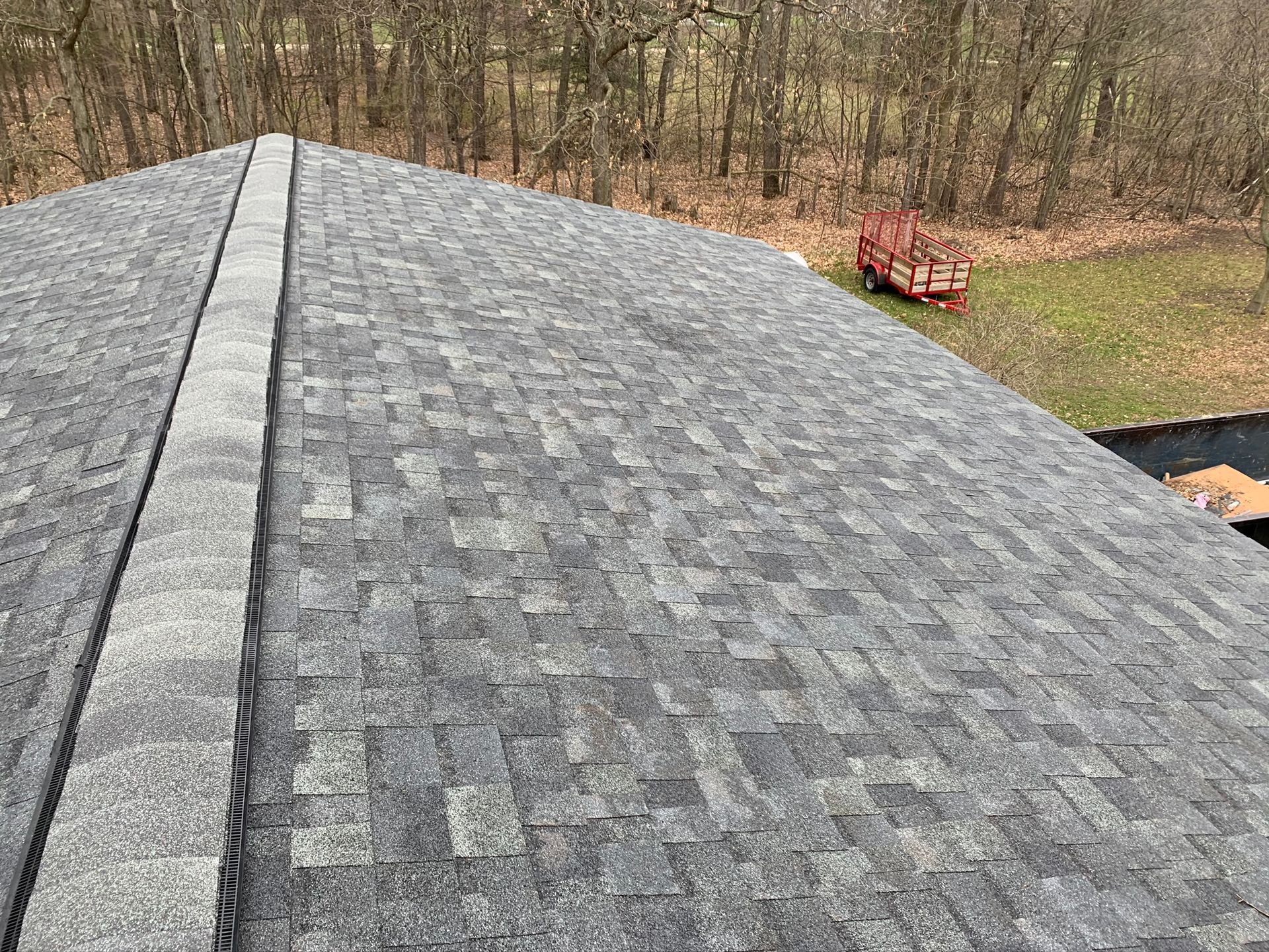 Gray shingled roof with ridge vent, overlooking a wooded yard and a red trailer below