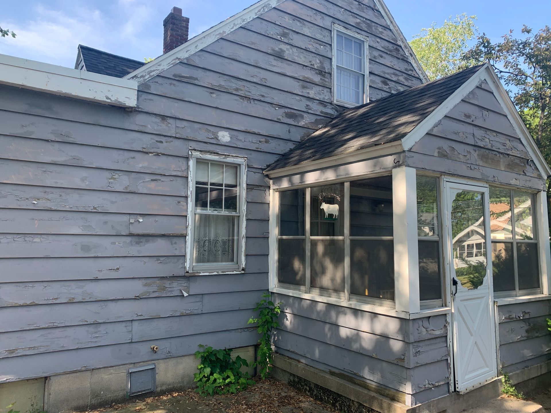 Gray weathered house exterior with a small enclosed porch and white-framed windows under a blue sky