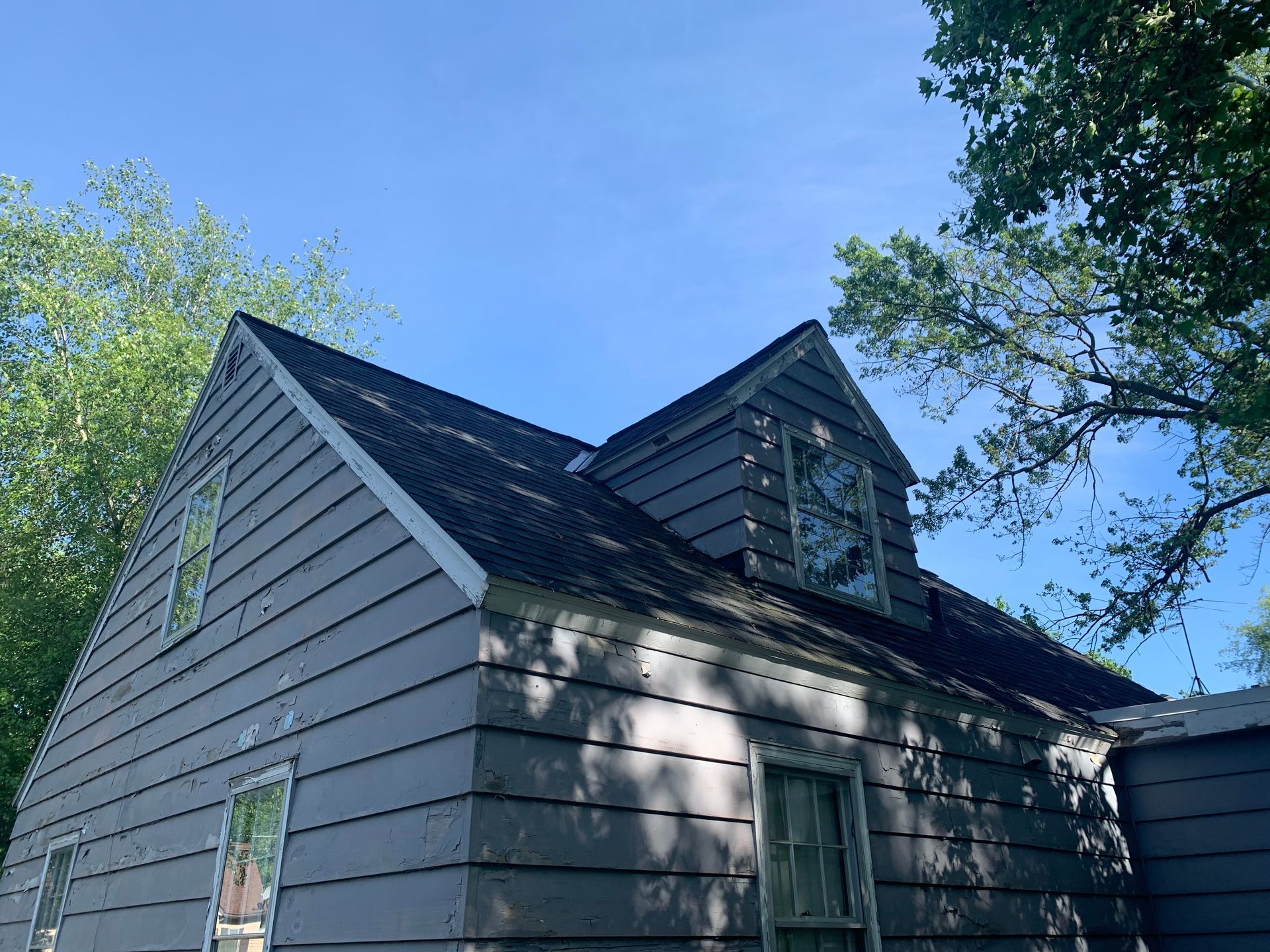 Gray house roof with dormer windows under a blue sky, surrounded by trees