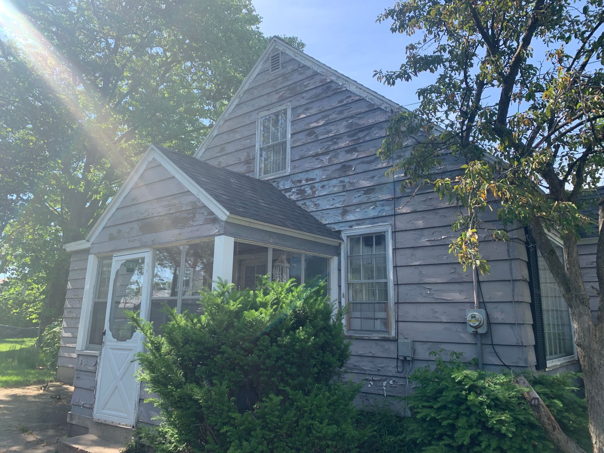 Weathered gray house with a small porch, surrounded by trees and bushes in bright sunlight