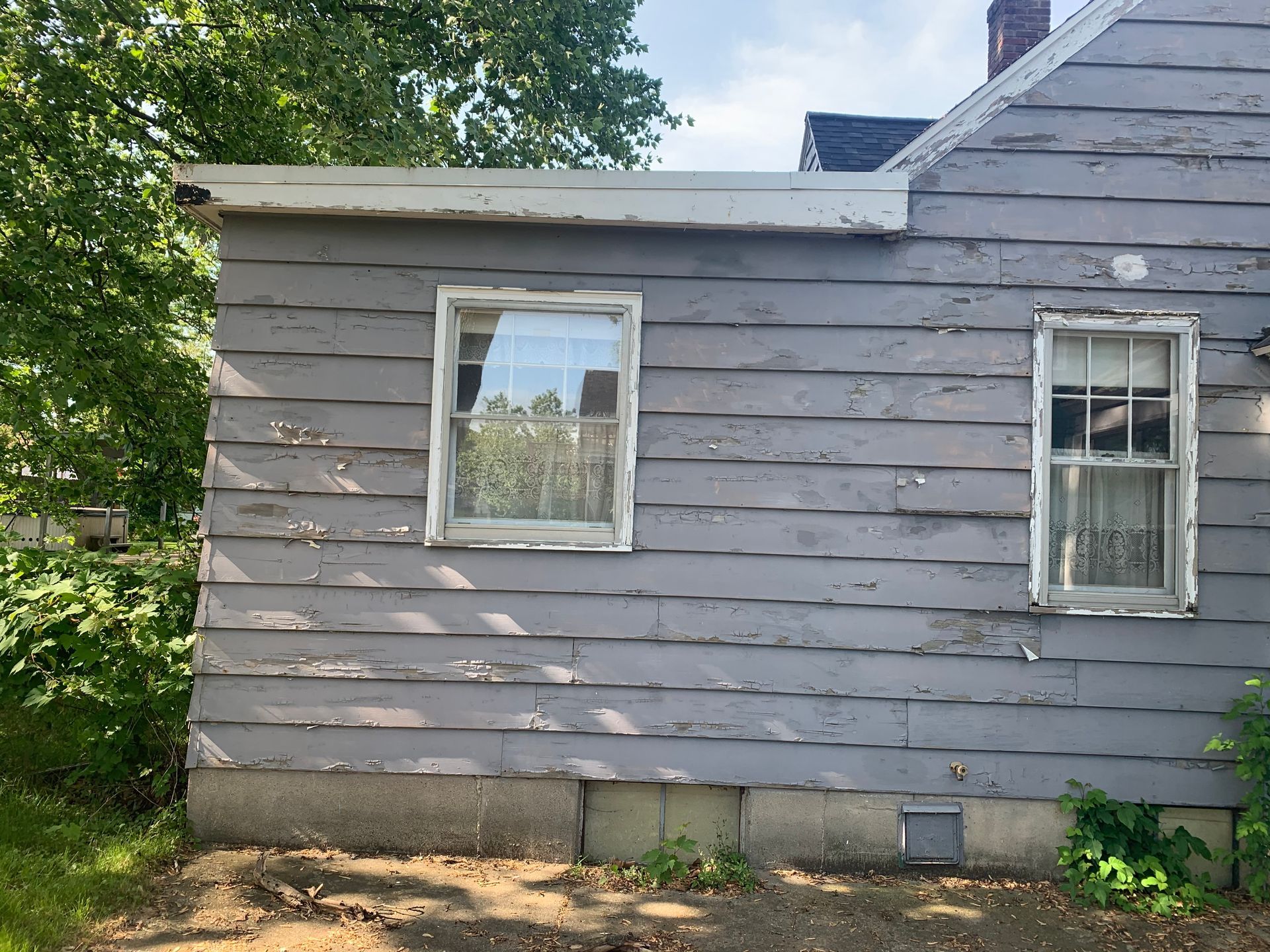 Gray house exterior wall with two windows and leafy trees at the left side