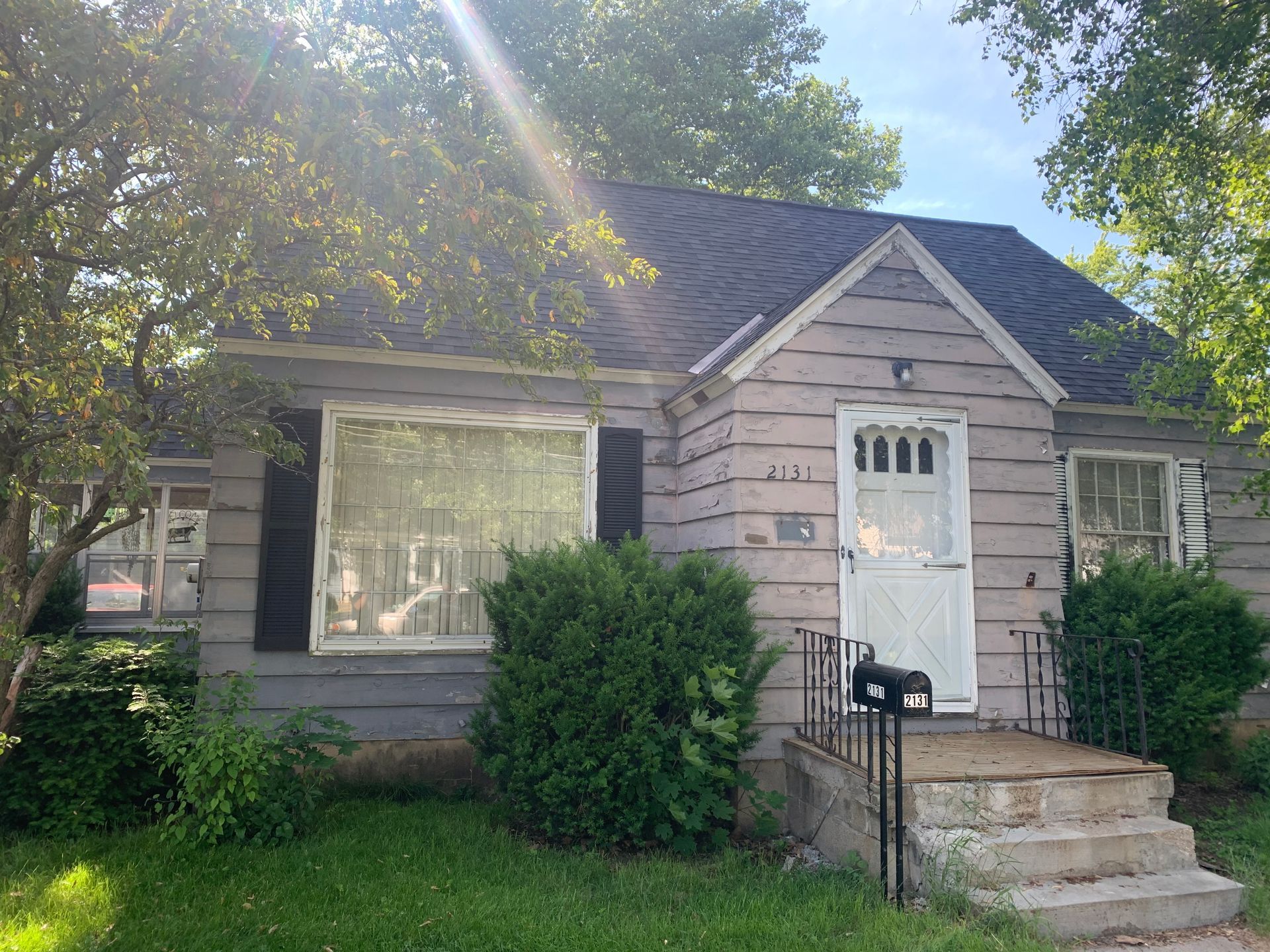 Small house with a white door, front steps, bushes, and sunlight through trees