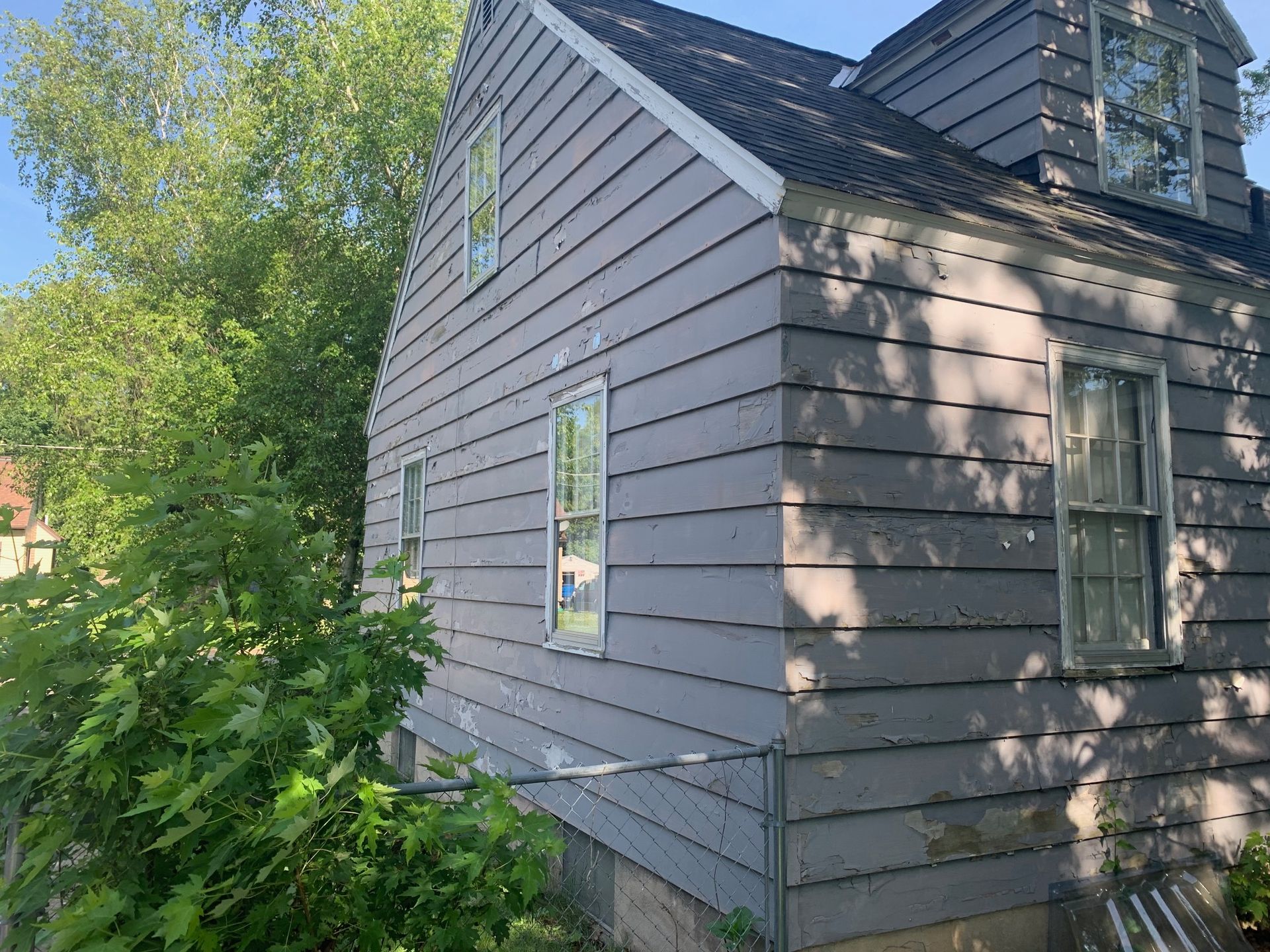 Gray house exterior with white-trimmed windows, shaded by trees and bushes.