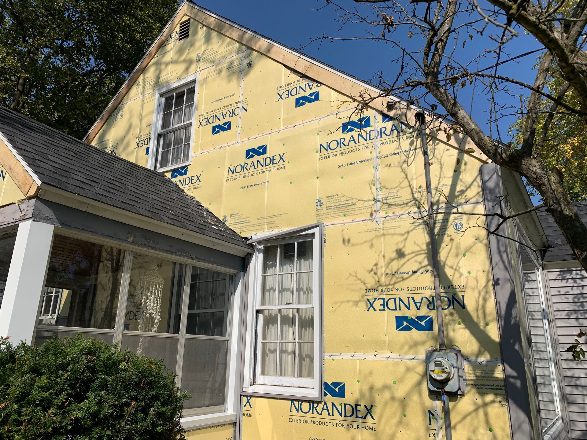 Yellow house under renovation with Tyvek wrap, white trim, and a tree in front