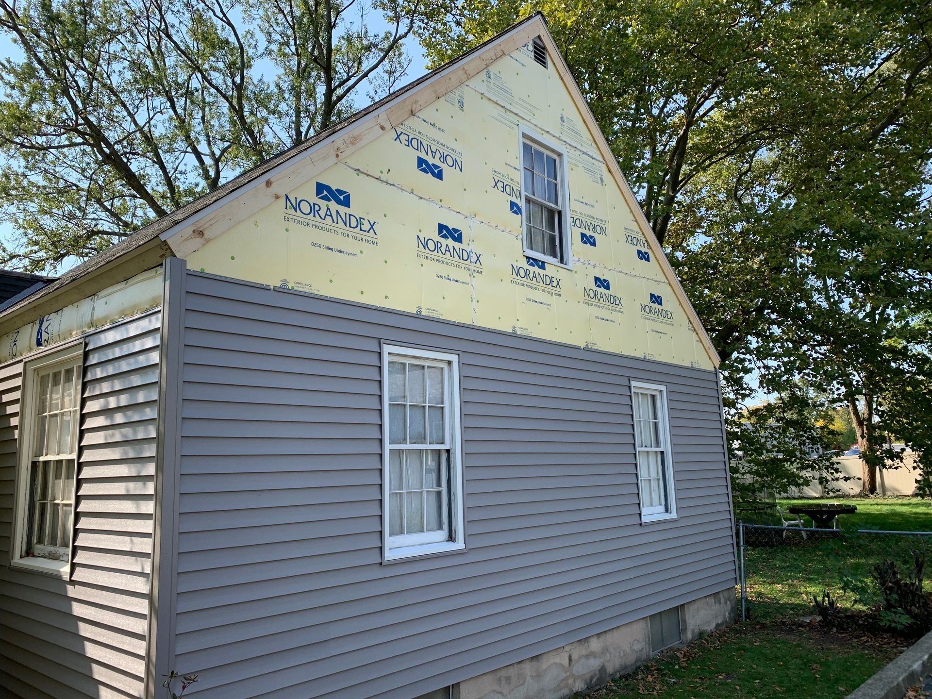 Side of a house under construction with gray siding and yellow sheathing, near trees and a yard.