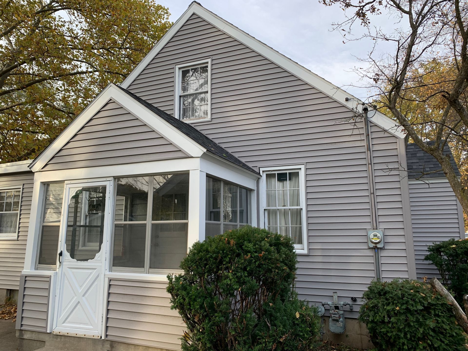 Gray house with white trim and enclosed front porch, surrounded by autumn trees and shrubs