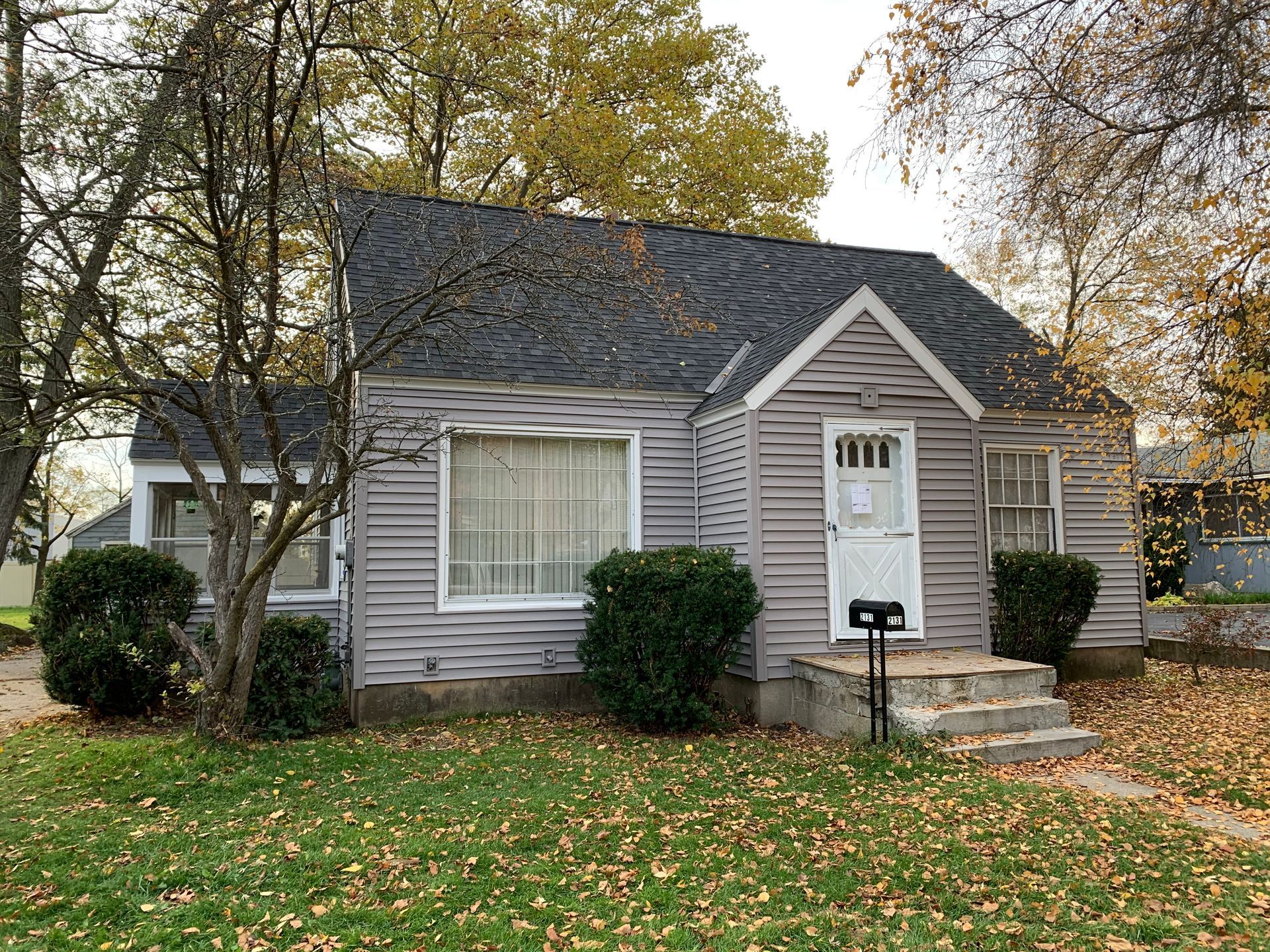 Small gray house with a front porch, white trim, and fallen autumn leaves in the yard.