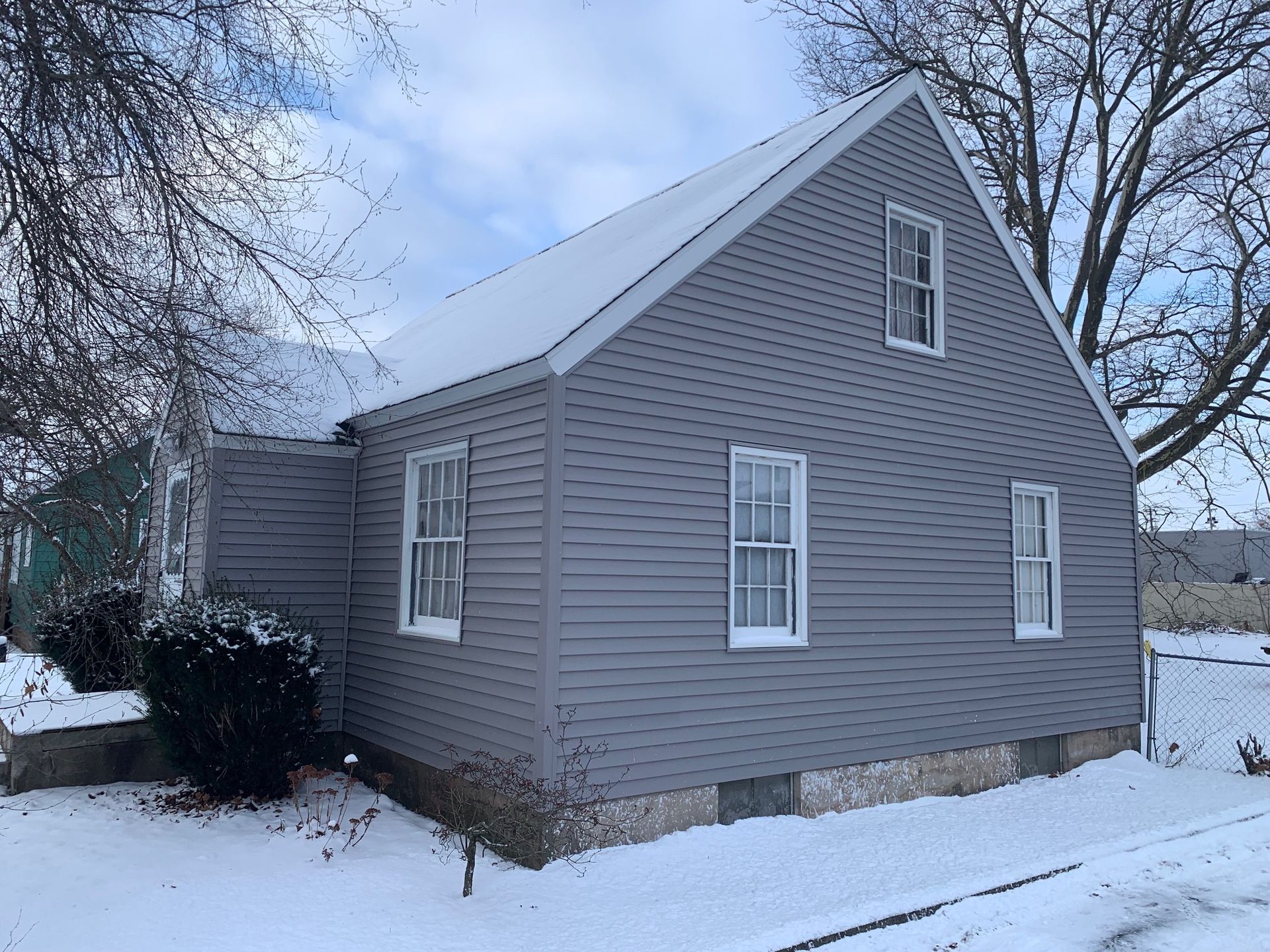 Gray house with a steep roof in a snowy yard, viewed from the side under a cloudy sky