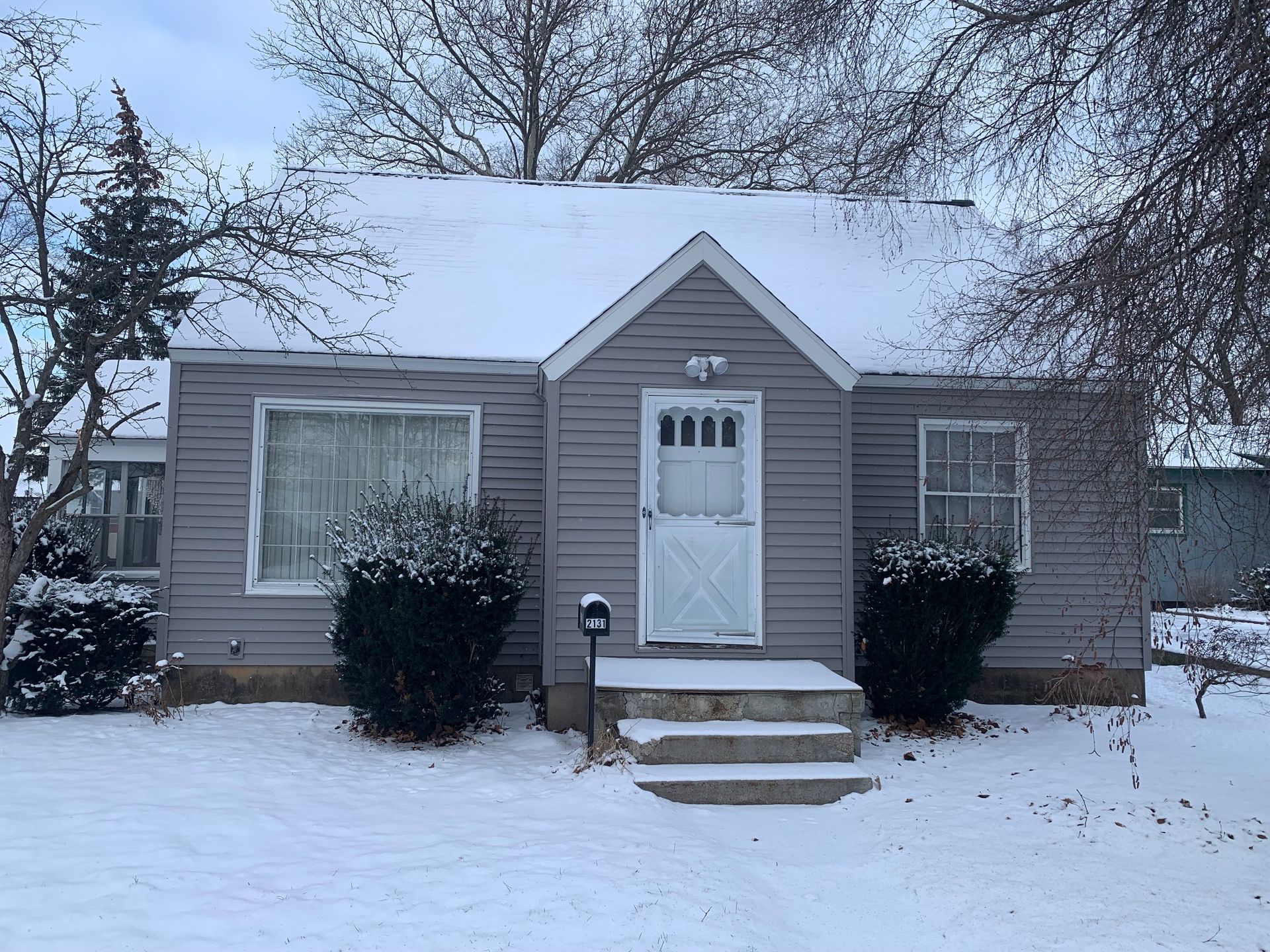 Snow-covered small house with a central front door, steps, and bushes in a winter yard