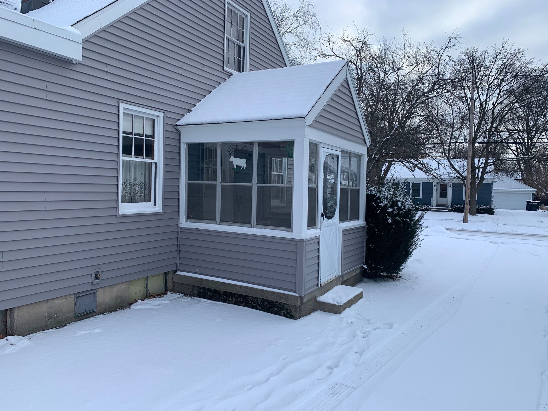 Snow-covered side yard with a small enclosed porch attached to a gray house.