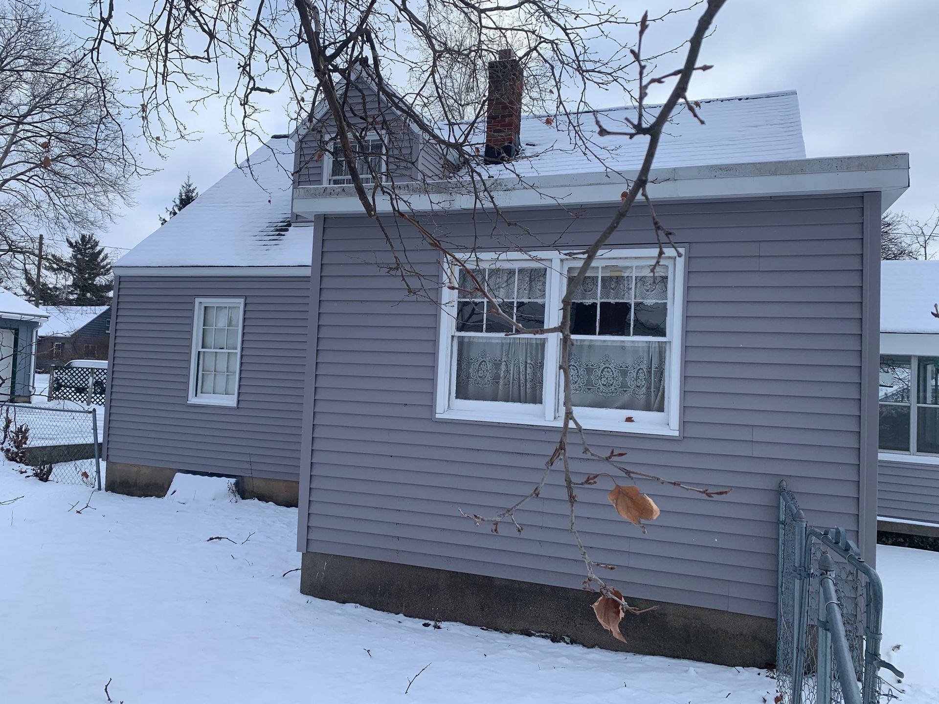Snow-covered gray house with a chimney and bare trees in a winter yard.