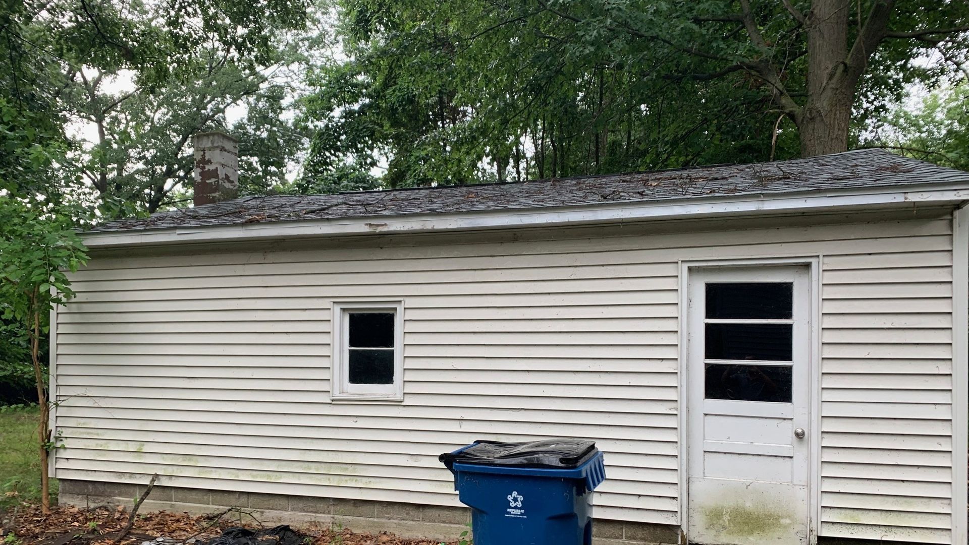 Small white shed with a blue trash bin in front, set among trees.
