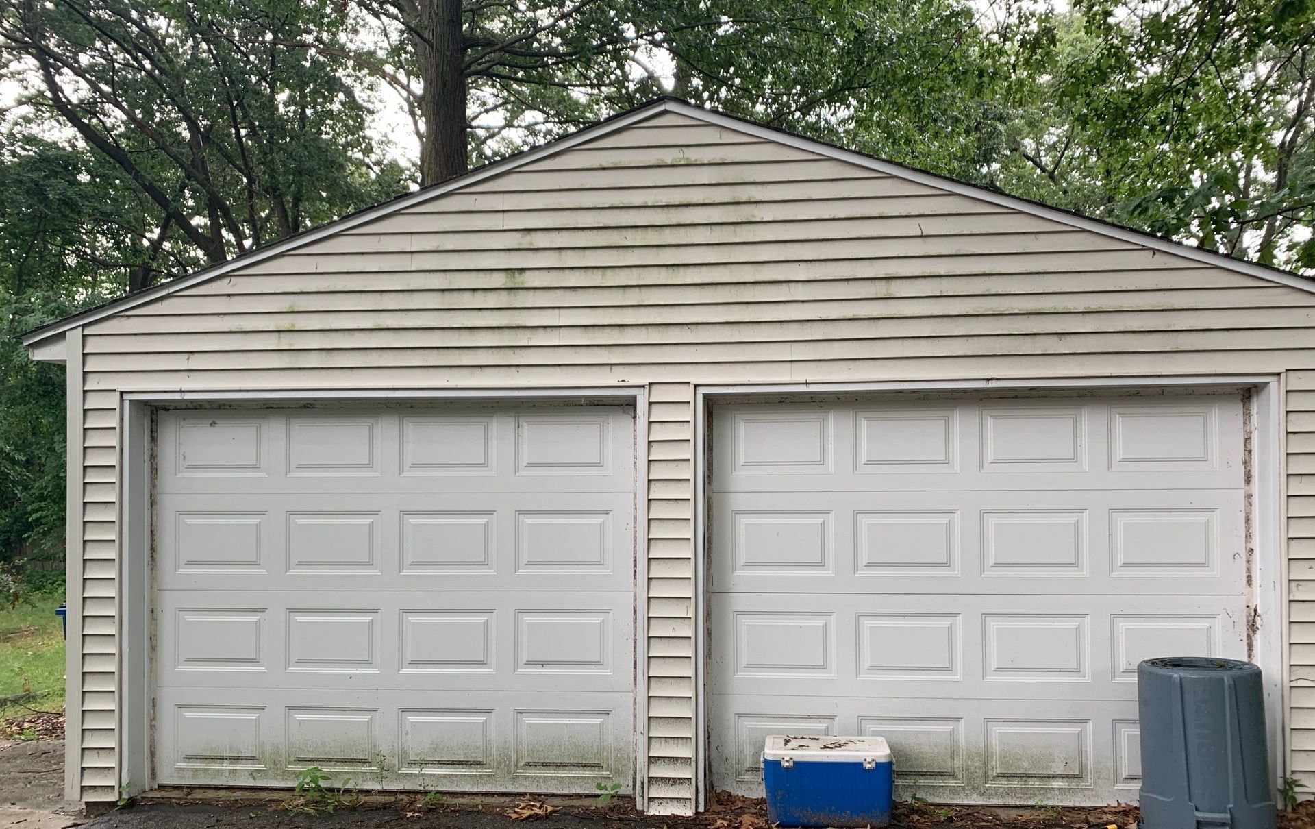 Two-car garage with white doors, beige siding, and trees in the background