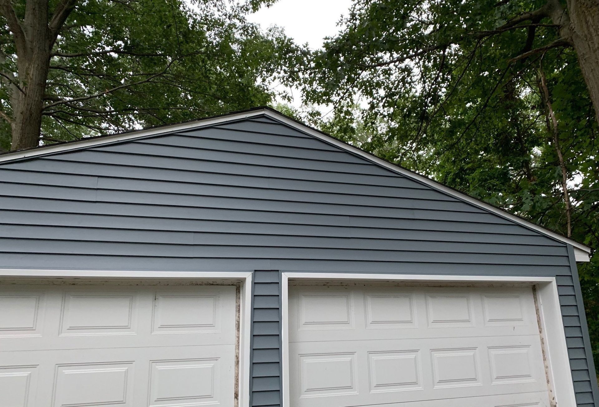 Gray garage with two white doors and a gabled roof under trees