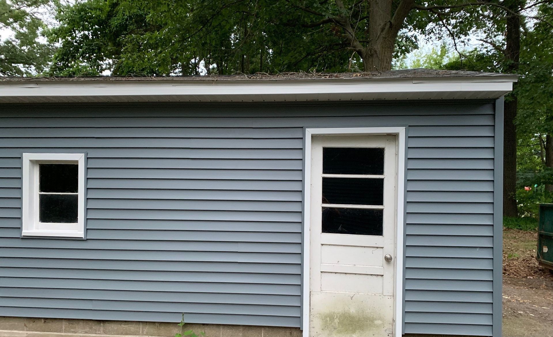 Small gray-blue shed with a white door and window in a wooded yard.