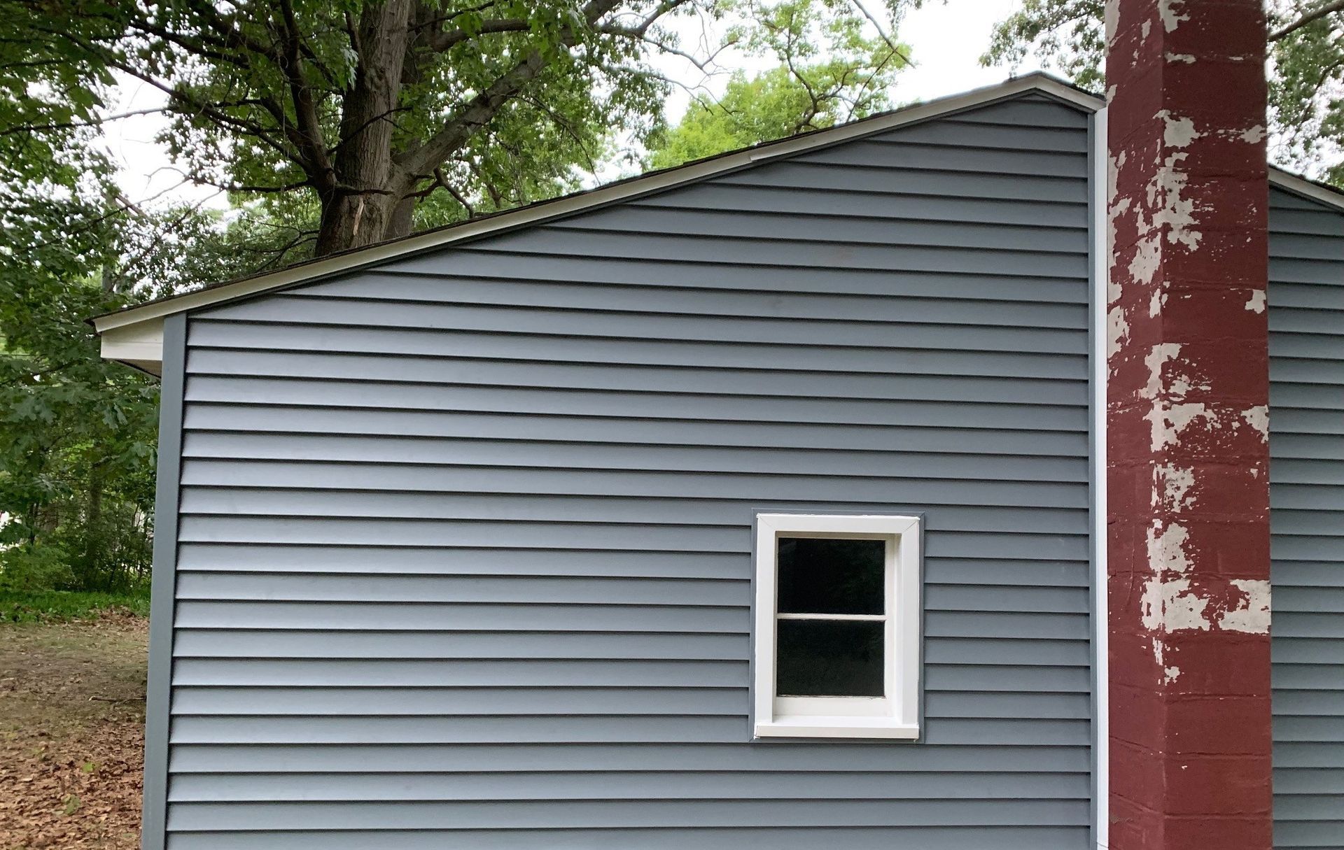 Gray-sided building with a small white-framed window and a red chimney against trees outdoors