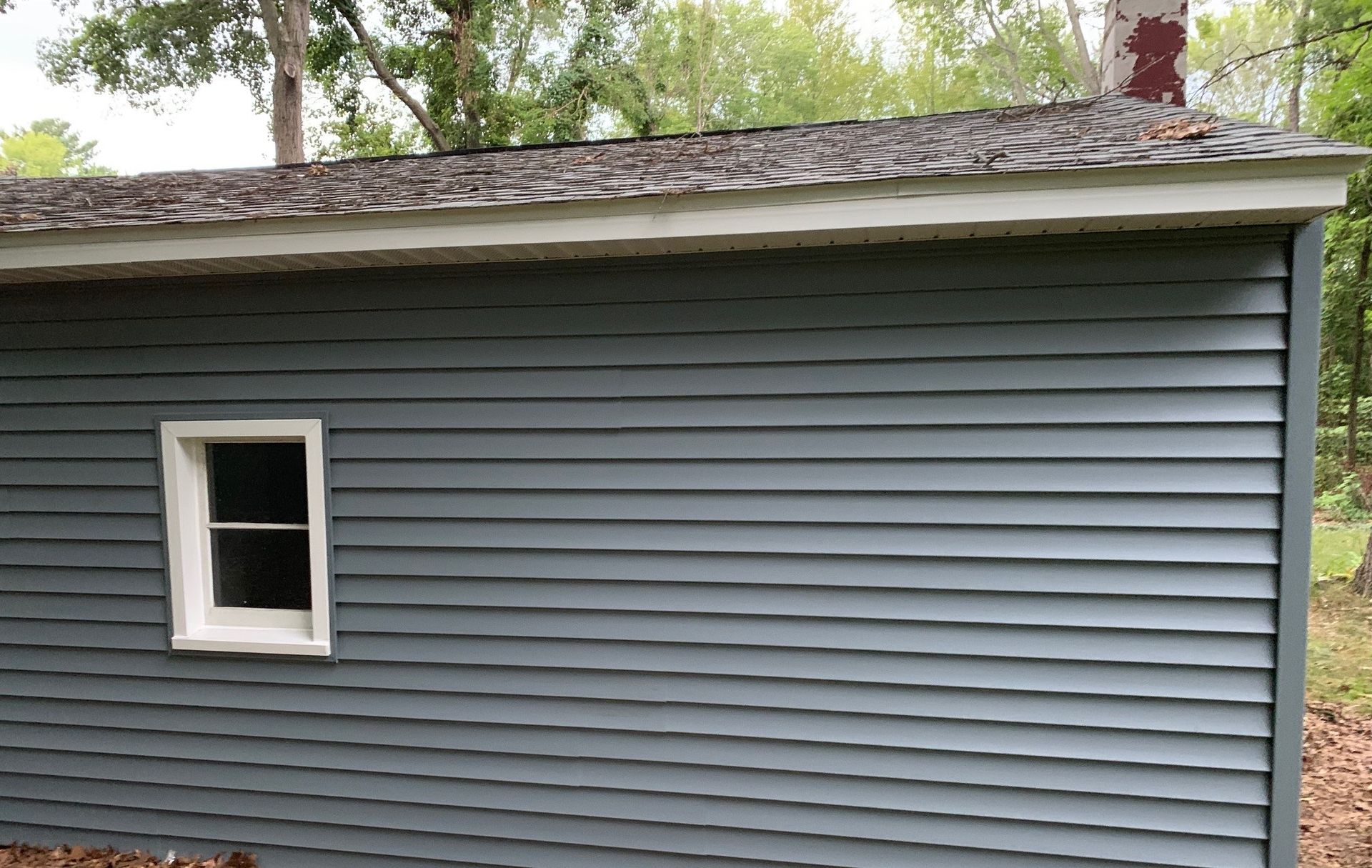 Gray house side with a small white-framed window and shingled roof under trees