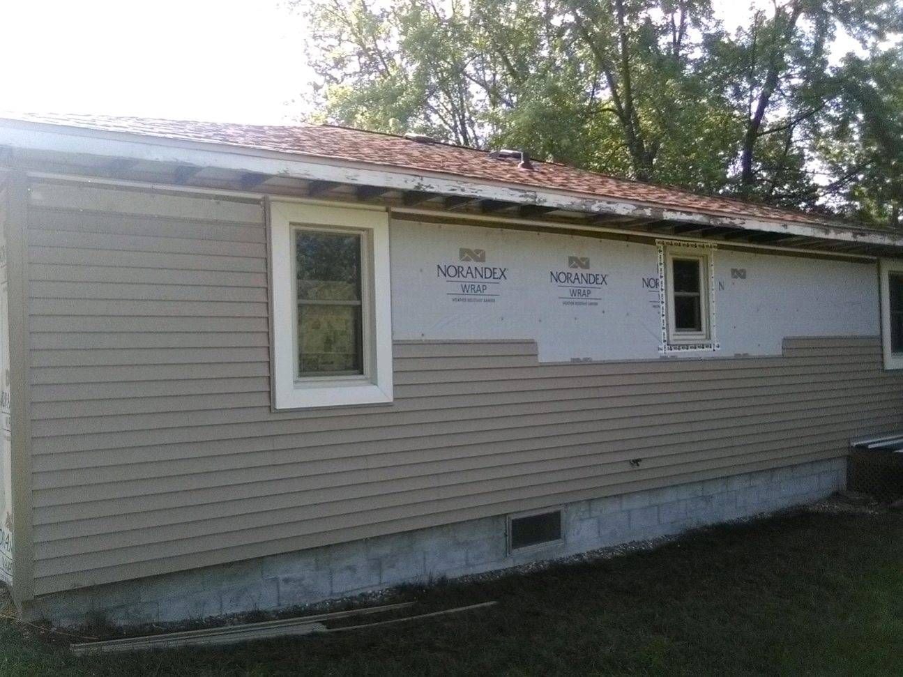 Siding-covered house exterior with a roof under repair and a white tarp over the wall.