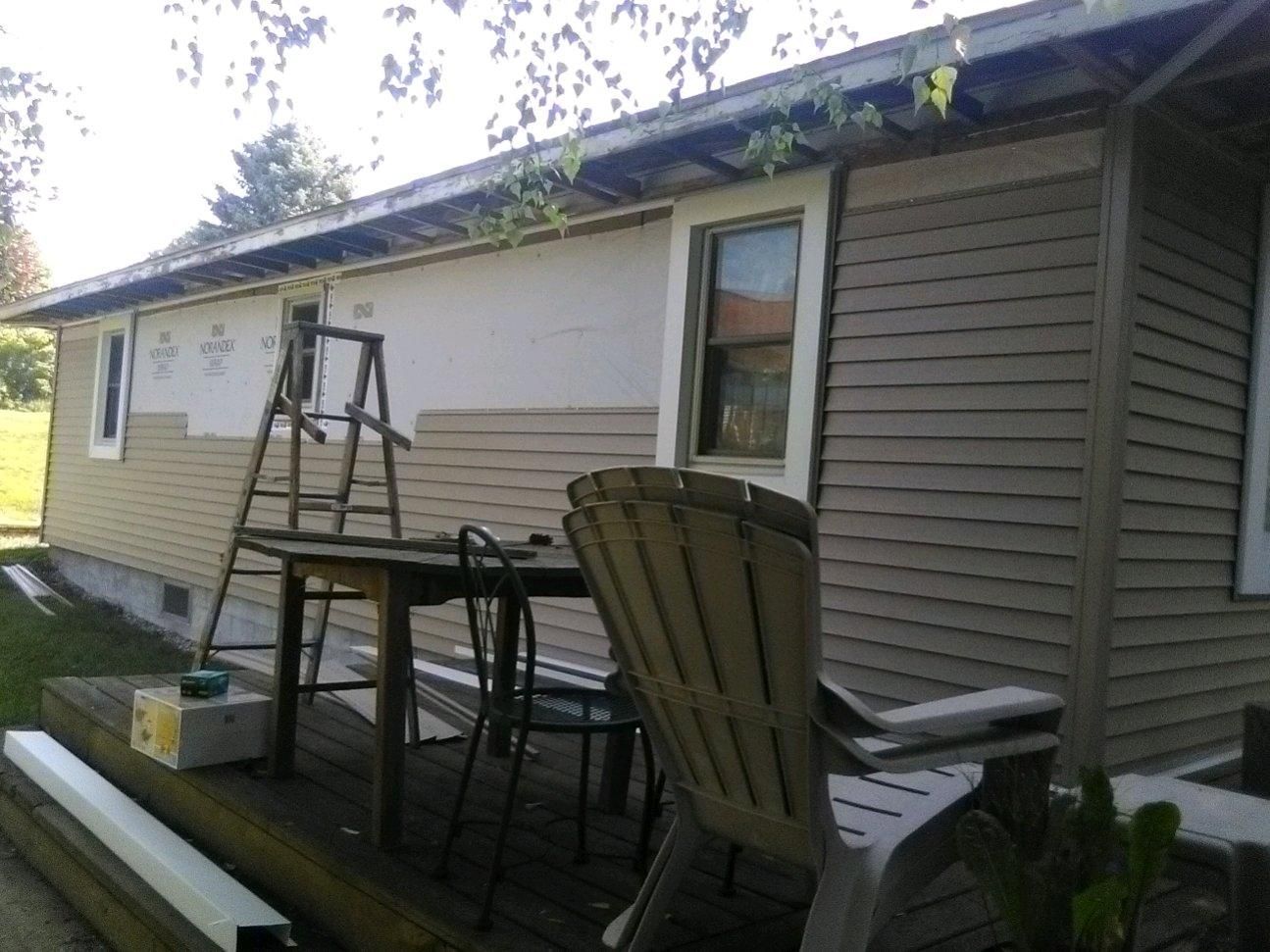 Side view of a house exterior with a small patio, chairs, ladder, and table under the eaves.