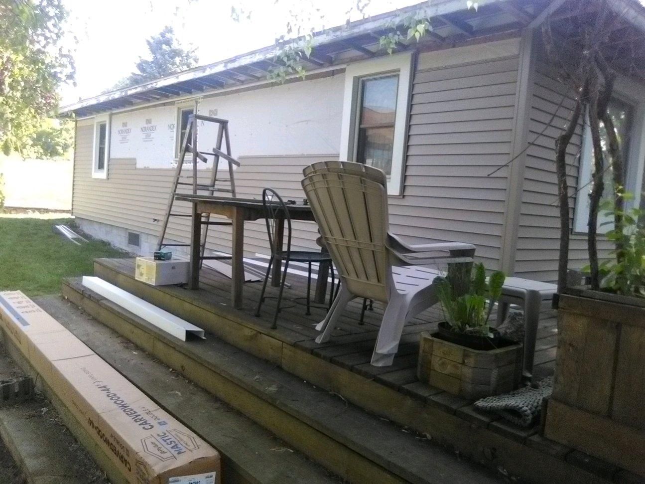 Back porch under construction with ladder, porch swing, chairs, and stacked lumber.