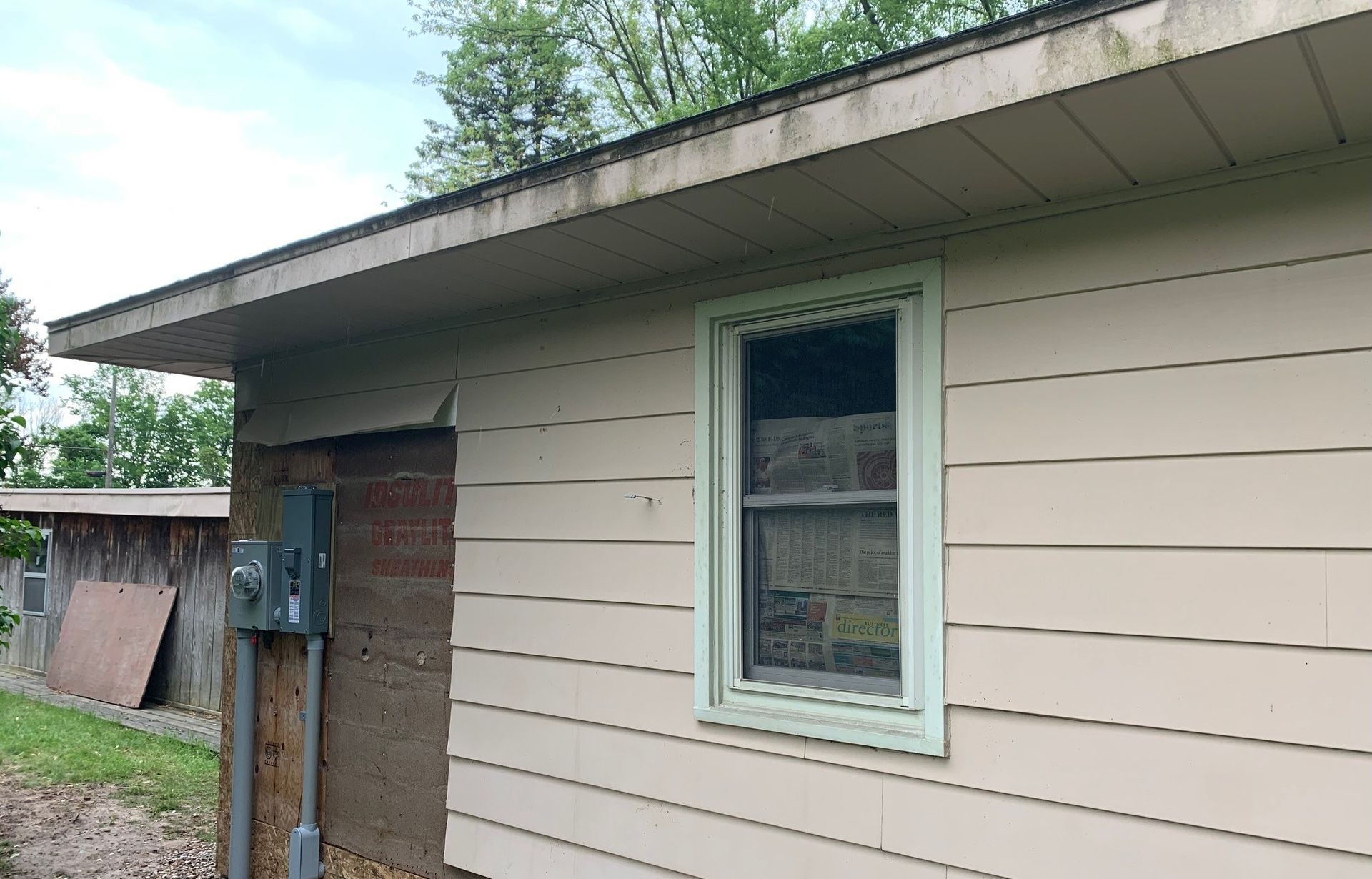 Side of a beige house with a window, utility boxes, and a wooden fence under an overcast sky