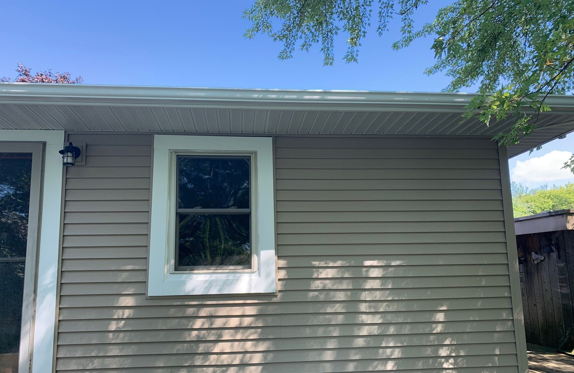 Exterior of a beige house wall with a white-trimmed window and roof eaves under tree shade