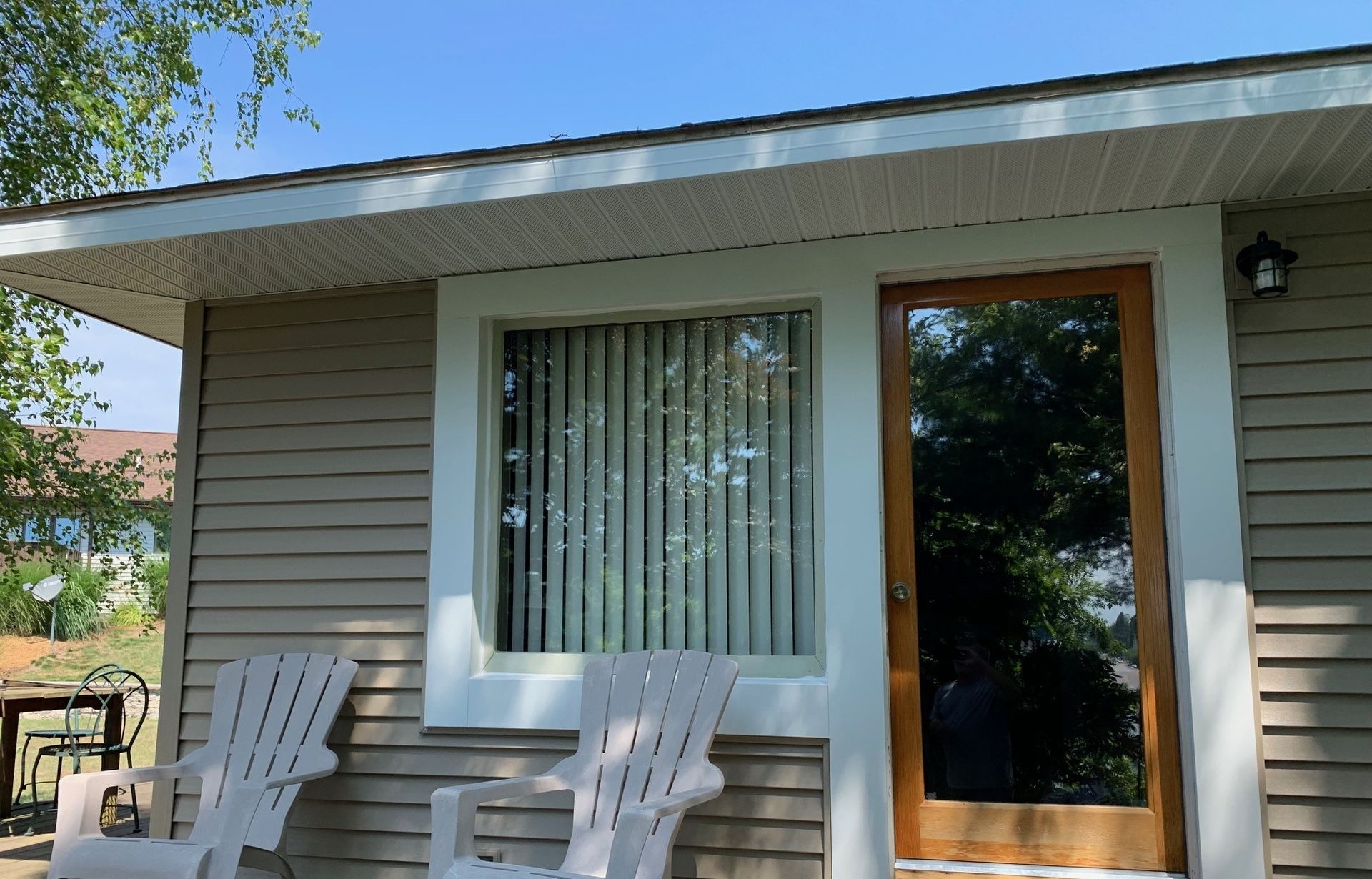 House porch with two white chairs, a large window, and a glass door under a sunny roof eave.