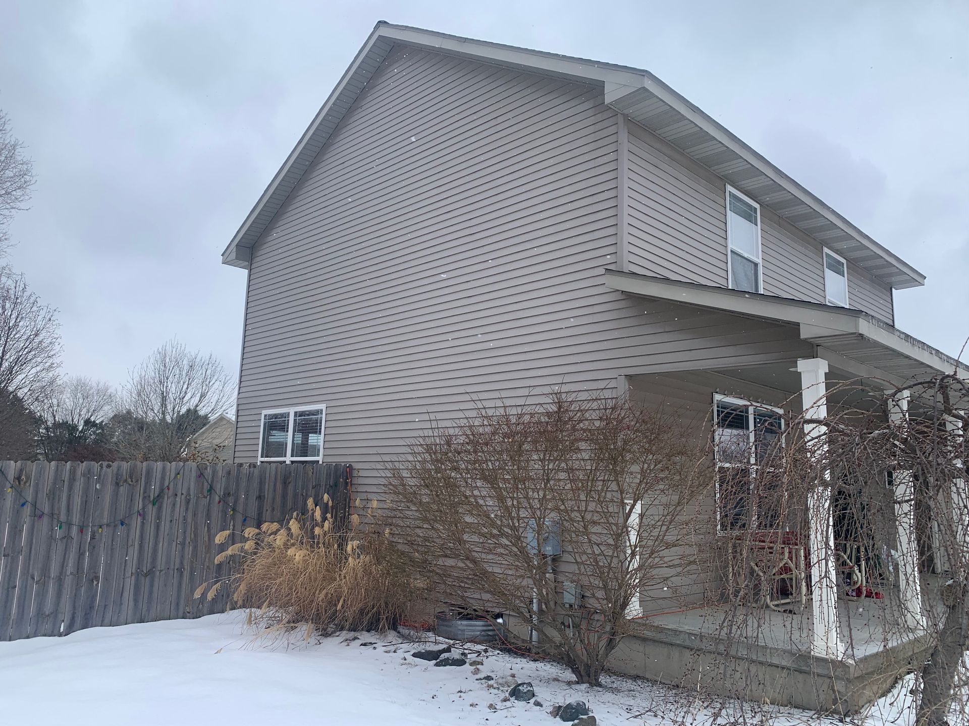 Snowy backyard beside a beige two-story house with a fenced yard and shrubs