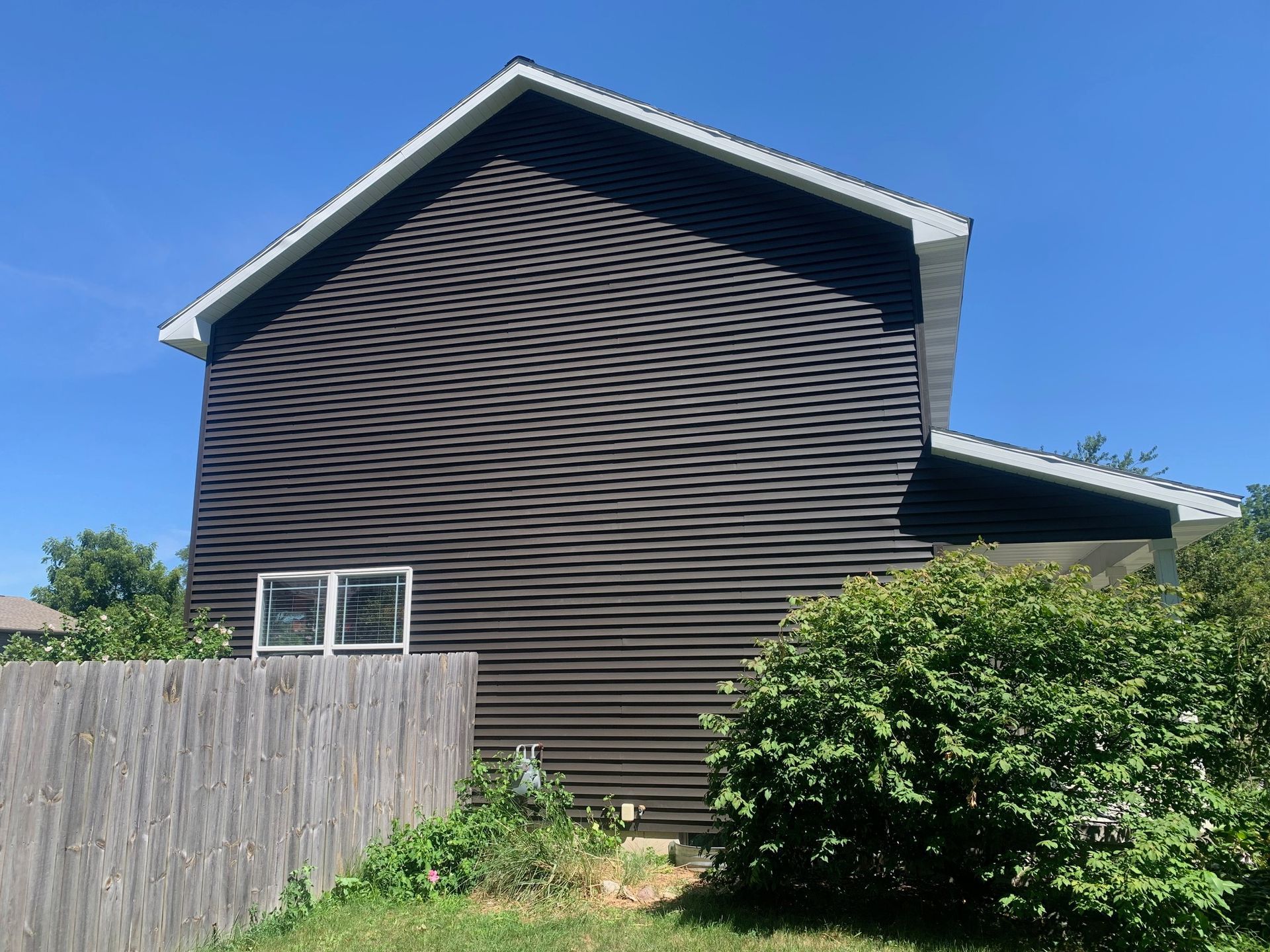 Dark-sided house with white-trimmed windows behind a wooden fence and green shrub under a blue sky
