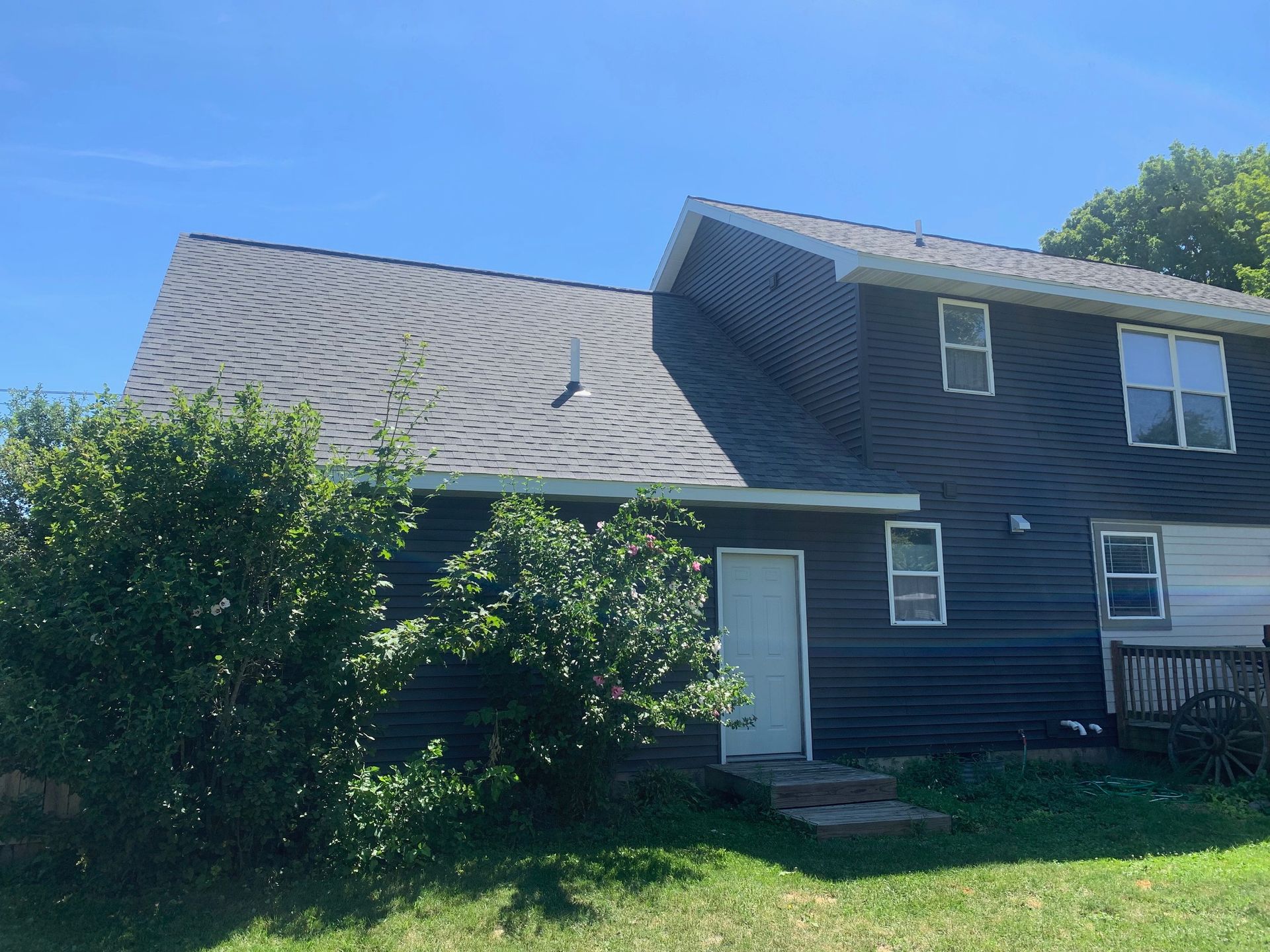 Two-story dark house with gray roof, white door, and bushes in front under a bright blue sky