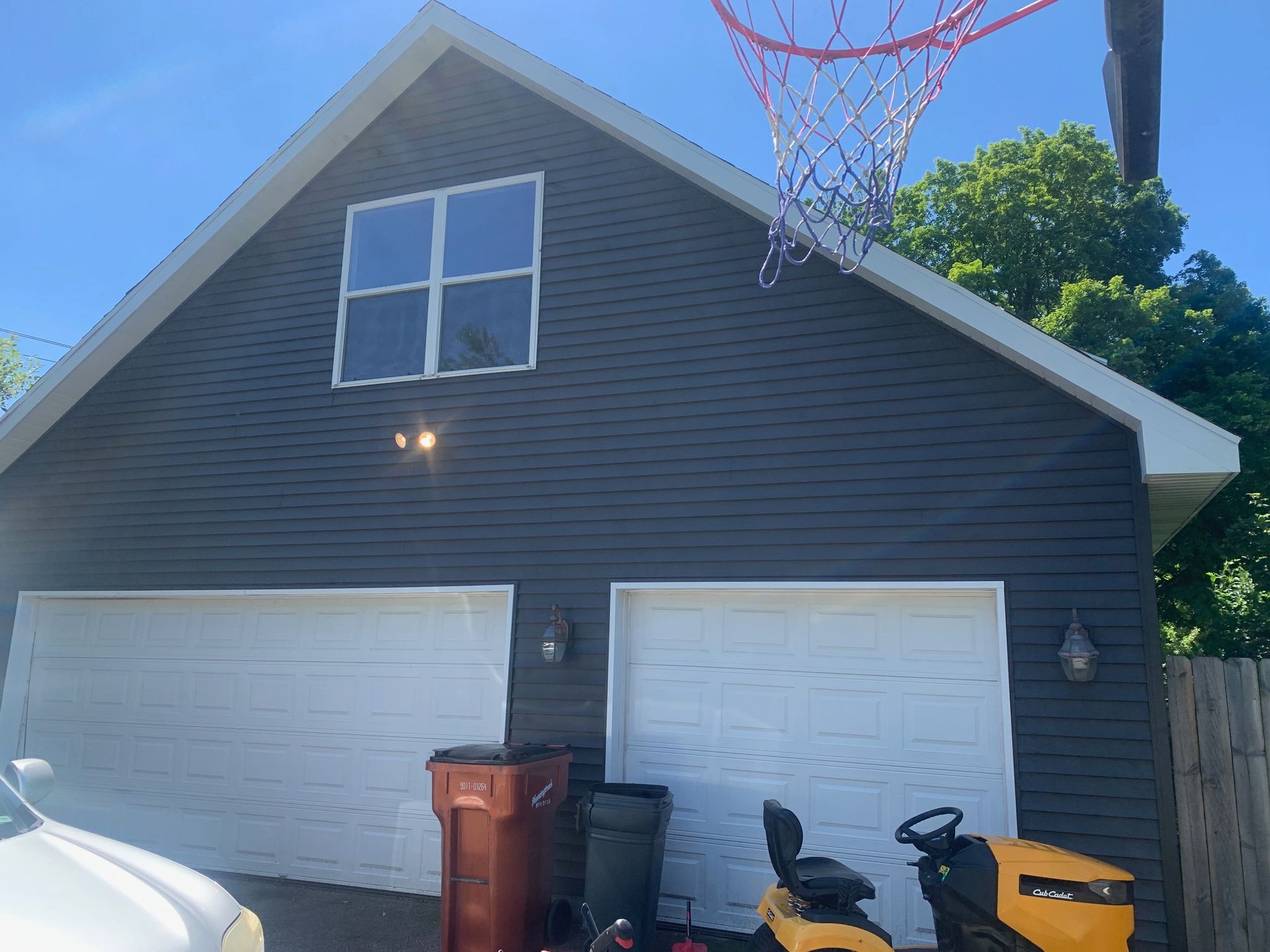 Suburban house with two white garage doors, a trash bin, and basketball hoop overhead in bright sunlight