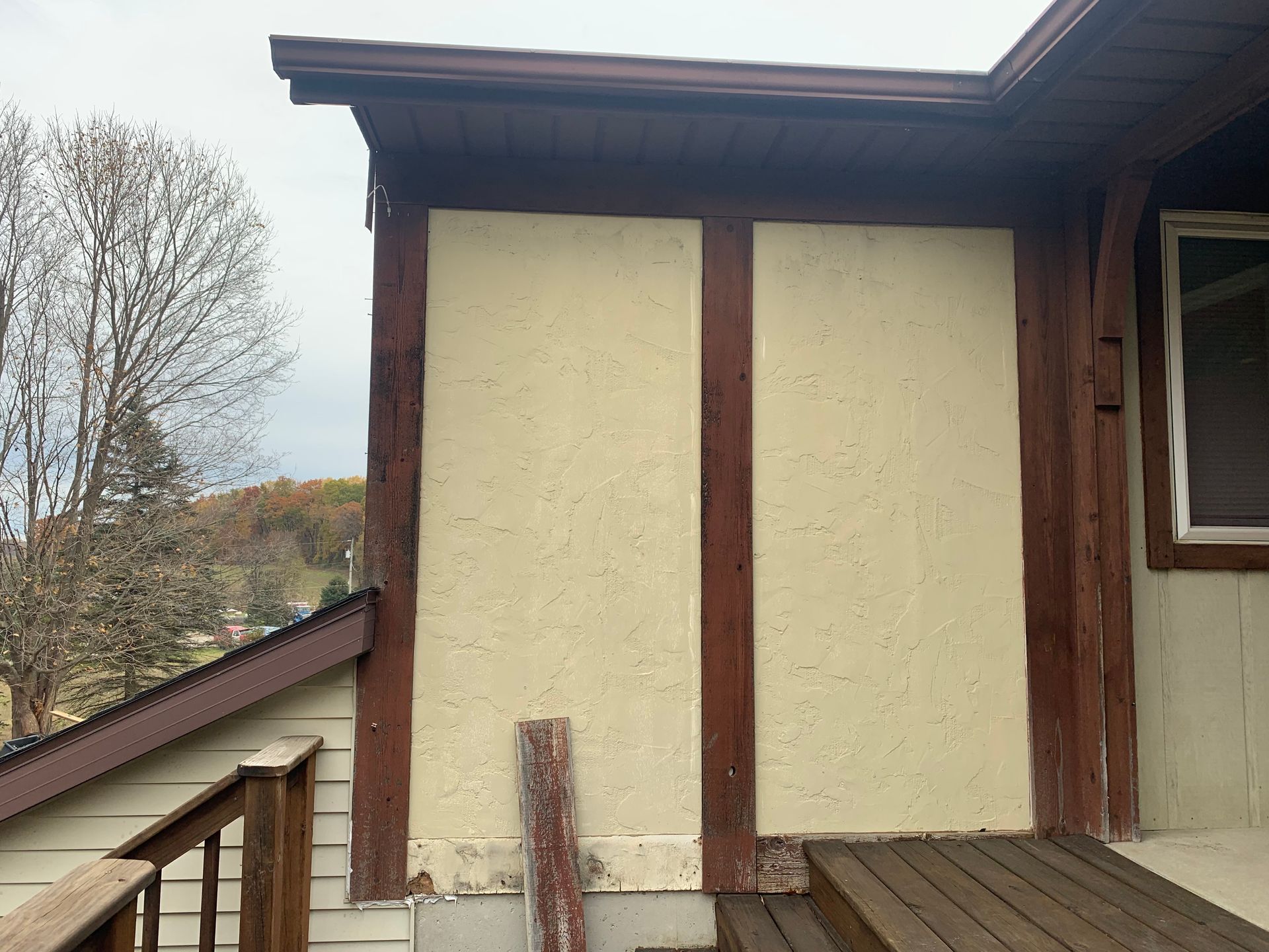 Exterior wall of a house with cream siding, brown trim, and a sloped roof under cloudy skies