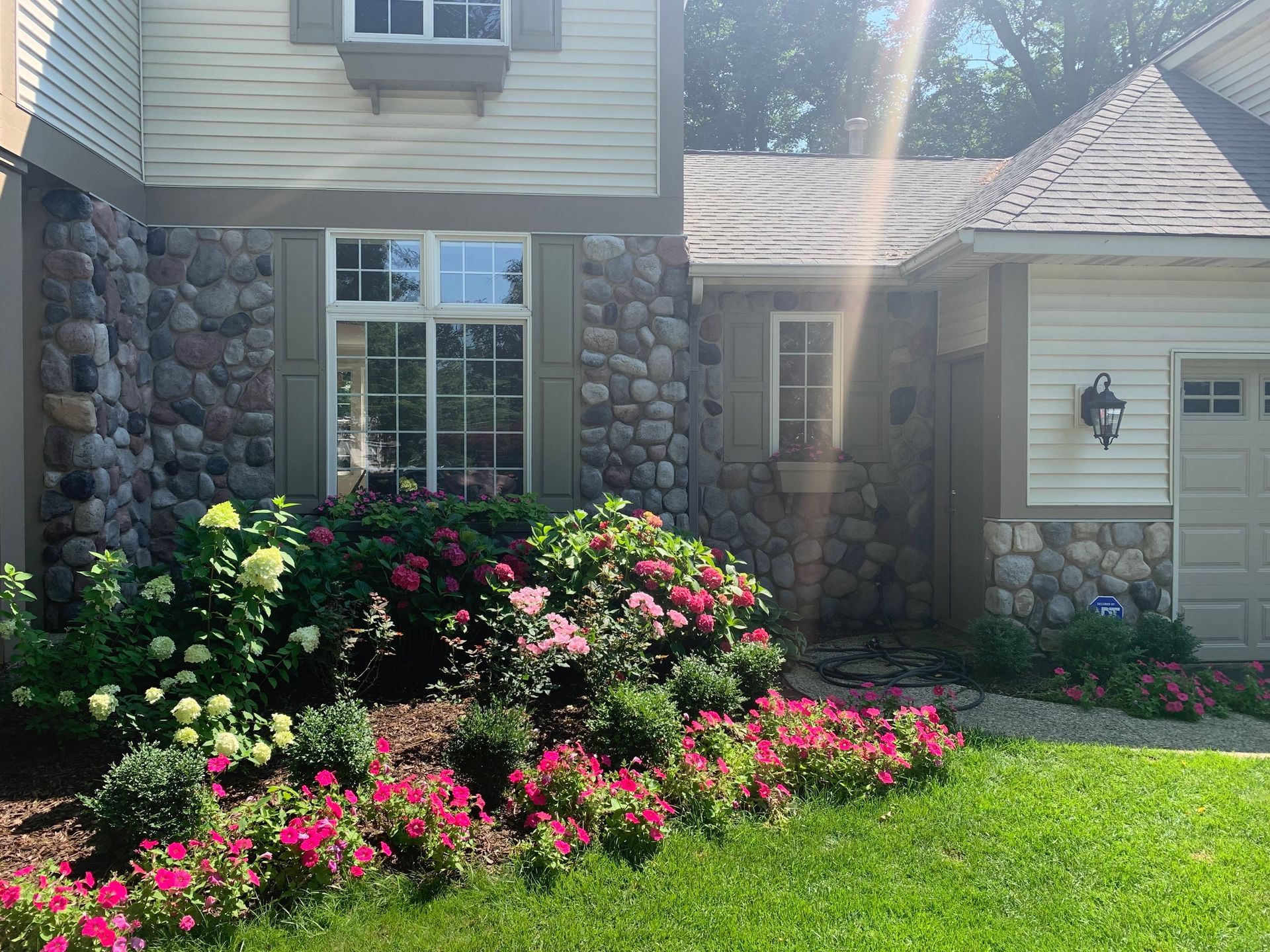 Front yard with pink and white flowers, green lawn, and a stone-and-beige house in sunlight
