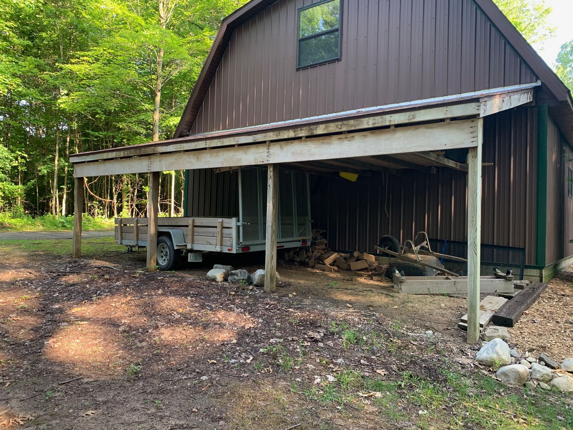 Brown barn with a covered porch, parked trailer, and stacked wood beside a wooded area.