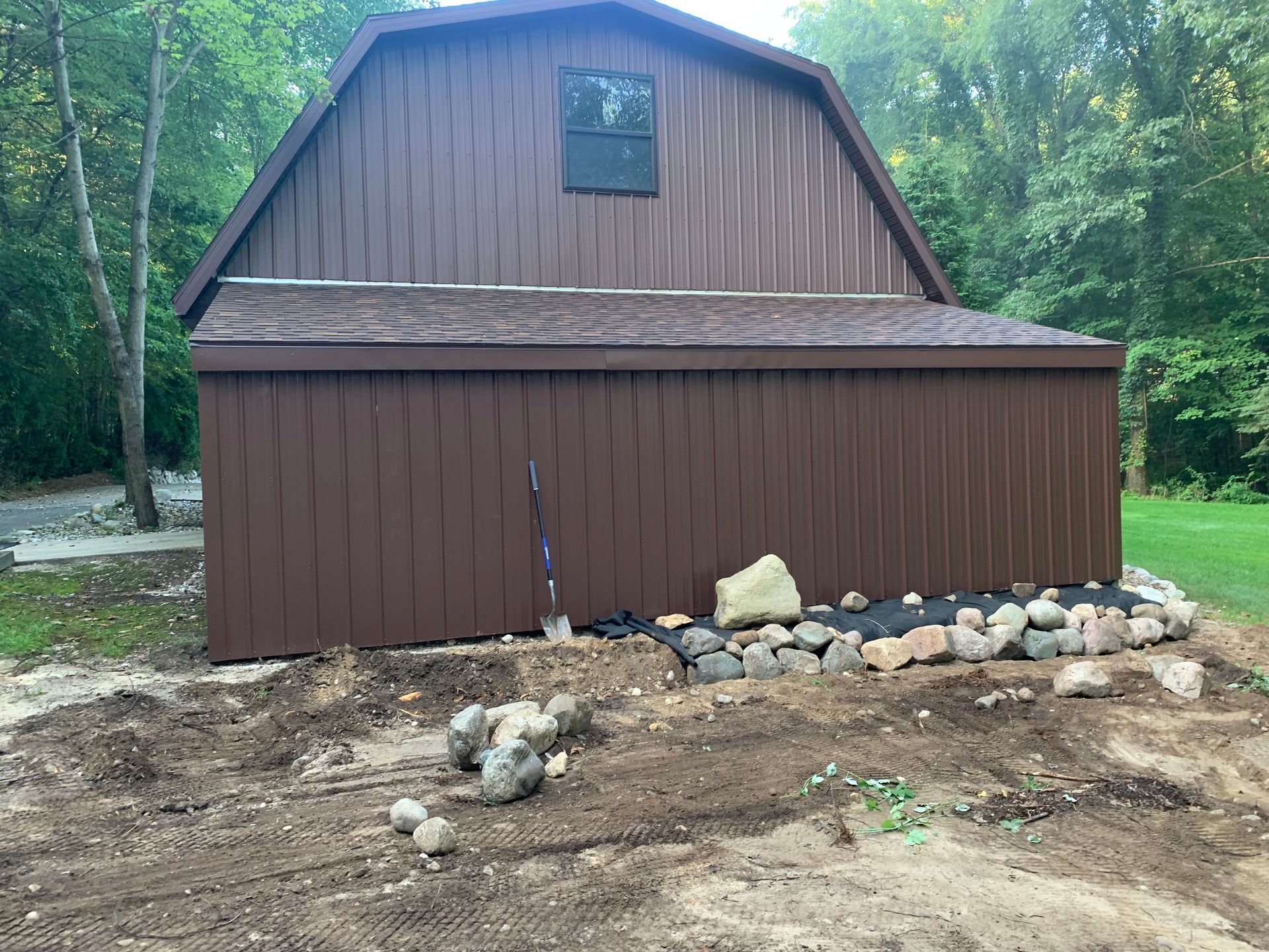 Brown barn-style shed beside a wooded area, with rocks and dirt in the foreground.