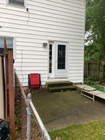 Back patio with concrete slab, white house door, red chair, and folding lounge chair near a fenced yard