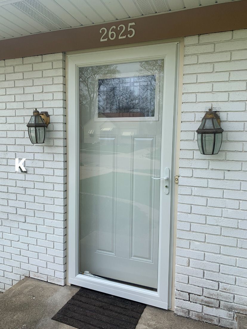 Front door with white storm door, house number 2625, brick wall, lantern lights, and black doormat