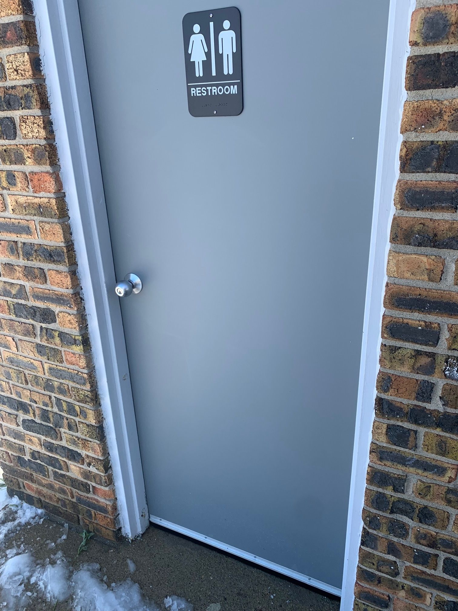Gray restroom door with a male/female restroom sign on a brick exterior wall.