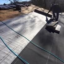 A worker installs black roofing material onto the wooden deck of a residential roof on a sunny day.