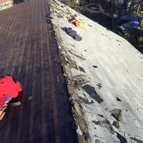 A person in a red jacket removes old shingles from one side of a residential roof while the other side remains covered.