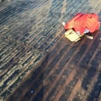A bright orange jacket and a small, light-colored bag lie on a dark, weathered wooden deck under sunlight.