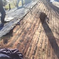 A person stands on a roof during a renovation, showing wooden planks exposed after old roofing material was removed.