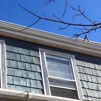 A view of gray shingled siding, a double-hung window, and white gutters against a clear blue sky with bare tree branches.