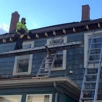 A worker in a high-visibility jacket stands on a ladder bridge extending between two ladders against a blue-sided house.