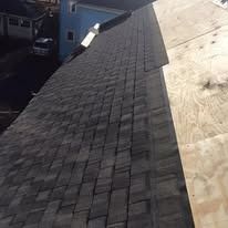 A sloped roof partially covered with dark, overlapping asphalt shingles, with a section of bare plywood decking.