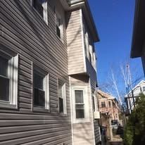 A side view of a house with light beige horizontal siding, multiple rectangular windows, and a sunny blue sky above.